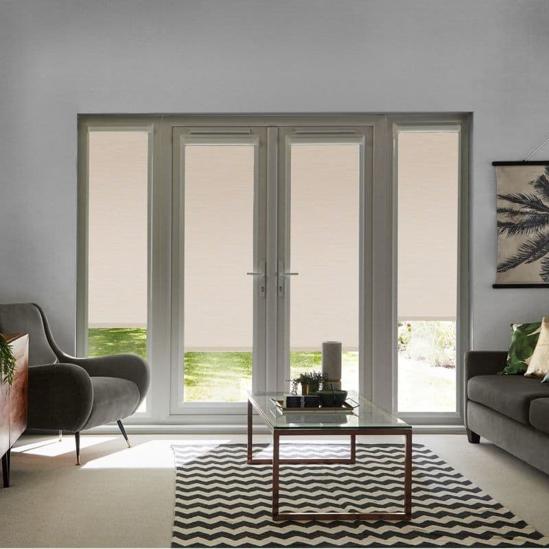 Beige roller blinds covering French doors and sidelights, lowered to mid-height revealing a narrow bottom view; filtering daylight into a neutral living room with armchair, sofa and glass coffee table.