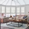 White pleated cellular shades span the bay windows and conservatory roof, lowered to diffuse daylight; cosy living room with beige sofa, chunky pink throw, wooden coffee table and blue rug.