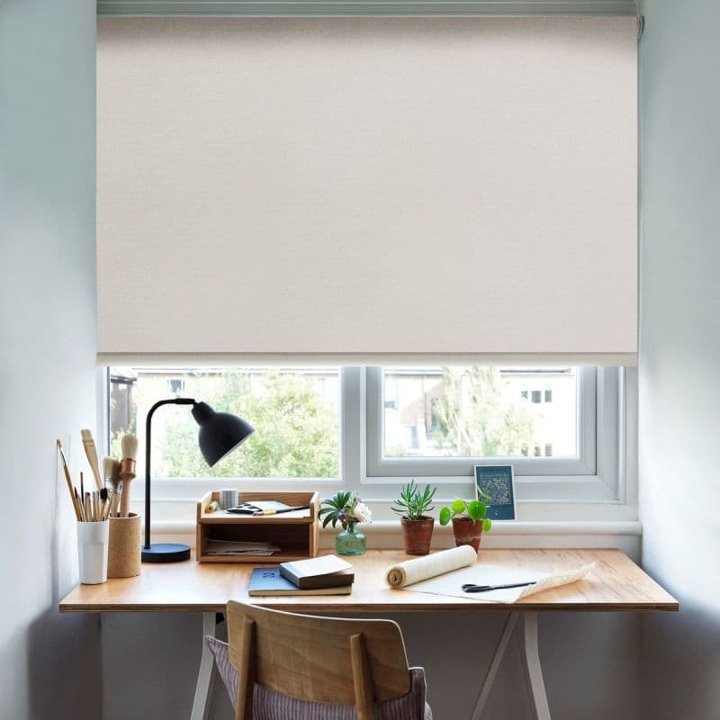 A cream roller blind lowered almost fully across the window, diffusing daylight; it covers a tidy wooden desk workspace with lamp, art brushes, notebooks and small potted plants, daytime view.
