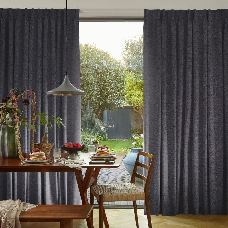 Thick pleated dark-gray curtains, hung and drawn aside to reveal a glass door, framing and filtering daylight; a dining table set with pancakes and pendant lamp overlooks a backyard.