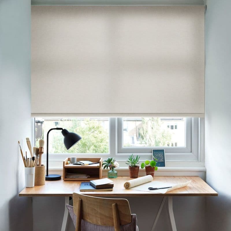 Beige fabric roller blind pulled down over a double window, filtering soft daylight. Below, a wooden desk with lamp, brushes, notebooks and small potted plants in a bright home studio.