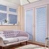 White horizontal Venetian blinds fitted inside a bay window and French doors, partially closed filtering sunlight; casting striped shadows across a cream sofa in a light-filled living room with rug.