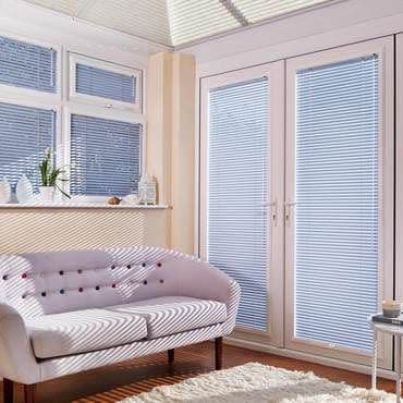 White horizontal Venetian blinds fitted inside a bay window and French doors, partially closed filtering sunlight; casting striped shadows across a cream sofa in a light-filled living room with rug.