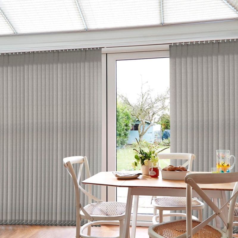Grey fabric vertical blinds span the patio doors, mostly closed but parted centrally to reveal a bright garden; a wooden dining table and cane-seated chairs sit in a sunlit conservatory.