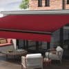 Red retractable awning with black support bar mounted above sliding glass doors, fully extended and shading an outdoor patio seating area with wooden table, chairs and brick façade in daylight.