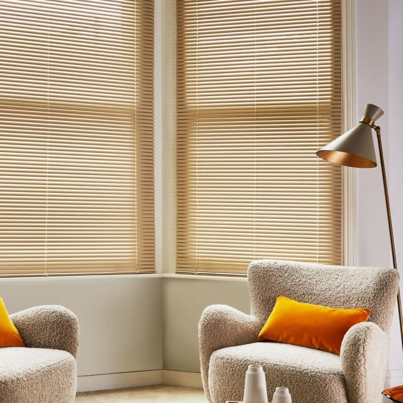 Horizontal beige venetian blinds covering bay windows, tilted closed to softly filter warm daylight; in a cozy living-room corner with textured boucle armchairs, orange cushions, a floor lamp, and neutral walls.