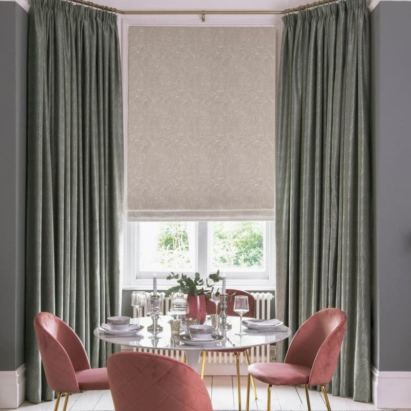 Green pleated, full-length curtains frame a lowered beige patterned Roman blind that filters daylight; in a dining room with a set table and pink upholstered chairs facing a garden view.
