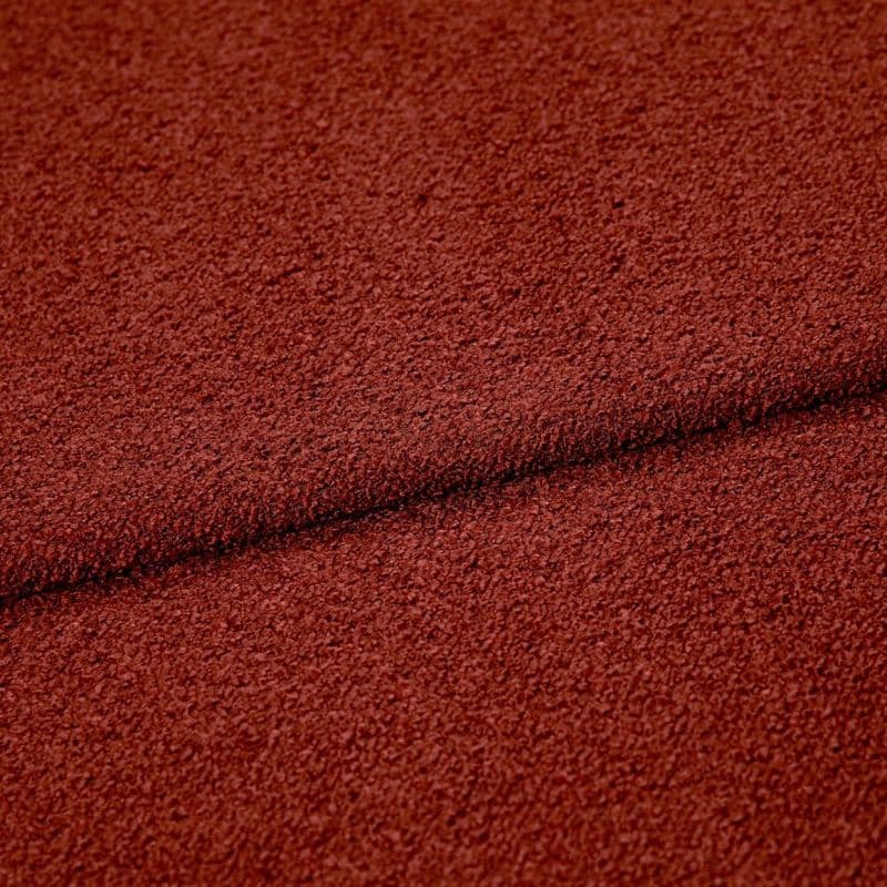 Rust-red textured boucle fabric folded across the frame, creating a dark seam; close-up studio view highlighting coarse looped surface and even, warm lighting.