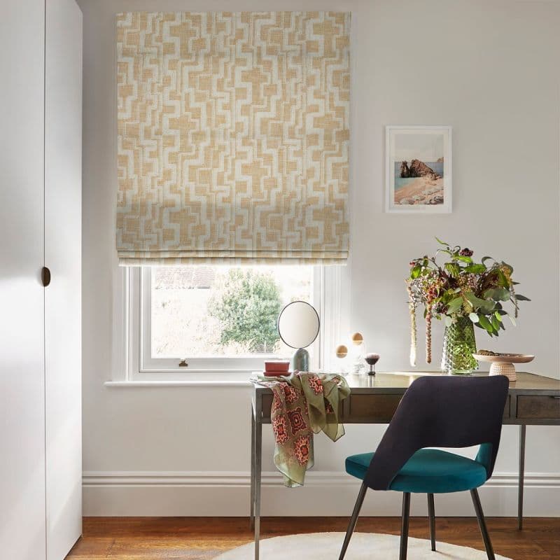 Object: beige-and-white patterned Roman blind, lowered over the window with geometric motif. Action: partially closed, filtering daylight through fabric. Context: small, bright home desk area with chair, vase of flowers.
