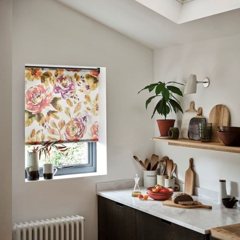 Floral roller blind lowered over a recessed window, printed with large watercolor roses, filtering soft daylight into a minimalist kitchen with marble countertop, wooden utensils, potted plant and cutting boards.