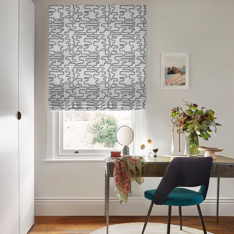 Roman blind with grey squiggle pattern, partially lowered and filtering daylight; over a window above a desk with vase of flowers, teal chair and framed print in a bright room.