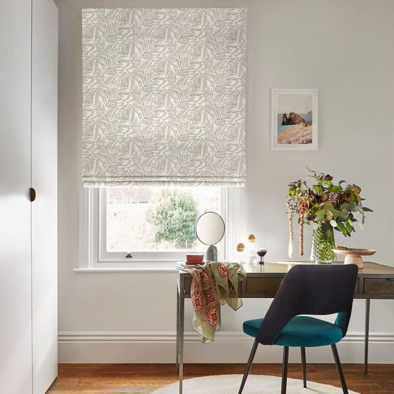 Floral-patterned Roman blind lowered over the sash window, pale leaf print, filtering soft daylight above a wooden desk with mirror, vase of flowers, teal chair and framed seascape.