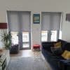 Two grey roller blinds lowered over adjacent French doors, filtering daylight and leaving bottom panes exposed; living room with dark sofa and yellow cushions, side table, plant, balcony chairs.