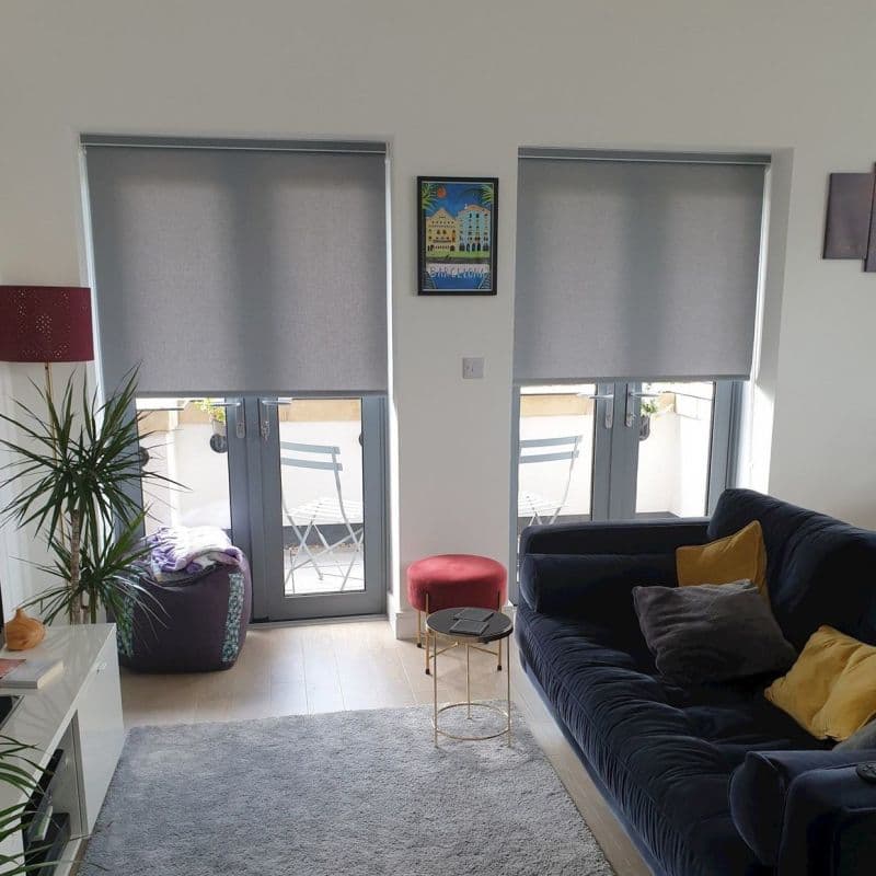 Two grey roller blinds lowered over adjacent French doors, filtering daylight and leaving bottom panes exposed; living room with dark sofa and yellow cushions, side table, plant, balcony chairs.