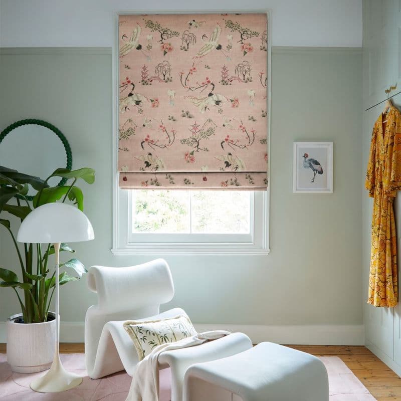 Pink Roman blind with crane motifs, partially lowered, filtering daylight into a pale-green sitting room with lounge chair, floor lamp, potted plant, framed bird print and hanging robe.
