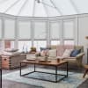 White honeycomb blinds covering tall conservatory windows and roof panels, lowered and closed, gently diffusing daylight; bright sitting area with beige sofa, knit throw, wooden coffee table, blue rug.