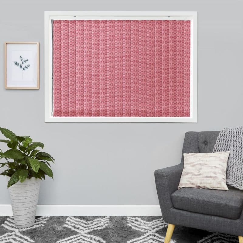 Pink patterned vertical fabric blinds, fully closed inside a white window frame, blocking light; in a gray living room with armchair, knitted throw, potted plant and framed botanical print.