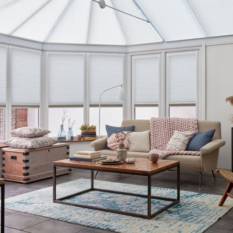 White cellular pleated blinds cover bay windows and roof panels, lowered to filter soft daylight; cozy living room with beige sofa, pink chunky throw, wooden coffee table and blue rug.