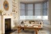 Grey layered zebra roller blinds covering three bay-window panels, partially lowered and filtering soft daylight; modern living room with beige sofa, rustic wooden coffee table, fireplace and neutral decor.
