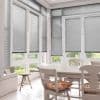 Horizontal aluminium Venetian blinds on tall French windows and doors, lowered with slats slightly tilted to filter daylight; sunlit dining nook with wooden table and chairs, patterned cushions, garden view.