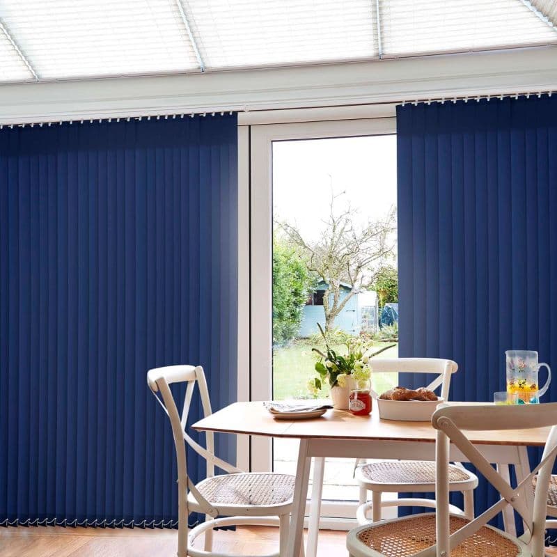 Navy blue vertical blinds over a sliding glass door, panels mostly closed with a central gap, filtering daylight while revealing a sunlit garden; dining table and chairs with vase and bowl.