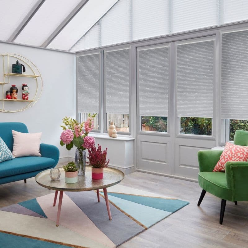 Light-grey pleated cellular blinds covering tall conservatory windows and glazed door, partially lowered and filtering daylight; bright sunroom with teal sofa, green armchair, round coffee table, geometric rug, floral arrangements.