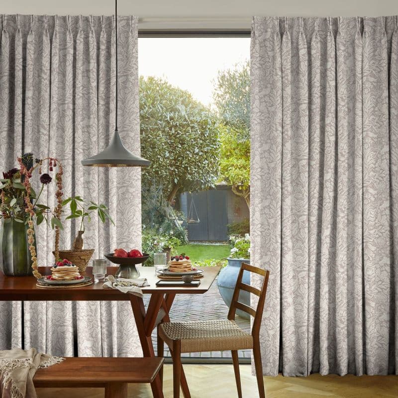 Pleated floral-patterned curtains, beige-gray and floor-length, drawn aside at a large glass patio door; revealing a sunlit dining table set for breakfast and a leafy backyard with trees and a shed.