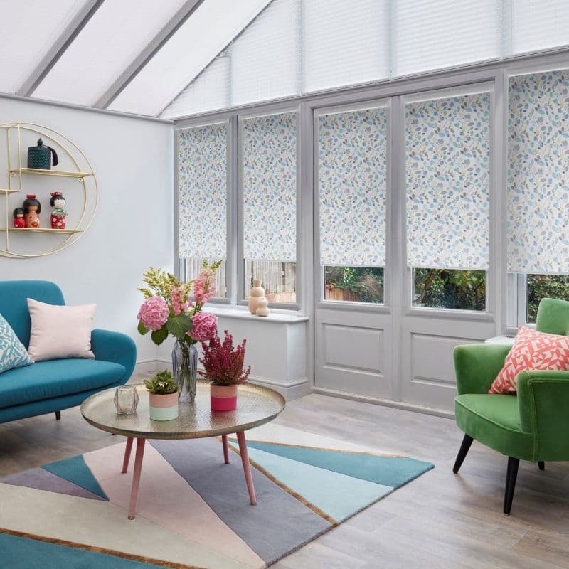 Floral-patterned roller blinds, lowered across tall conservatory windows and doors with a roof blind above, filtering soft daylight; bright sunroom with teal sofa, green armchair, coffee table and garden view.