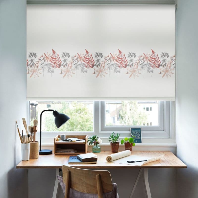 A white roller blind with a horizontal band of red and gray botanical prints, lowered across the window and filtering daylight above a wood desk with lamp, brushes, potted plants.