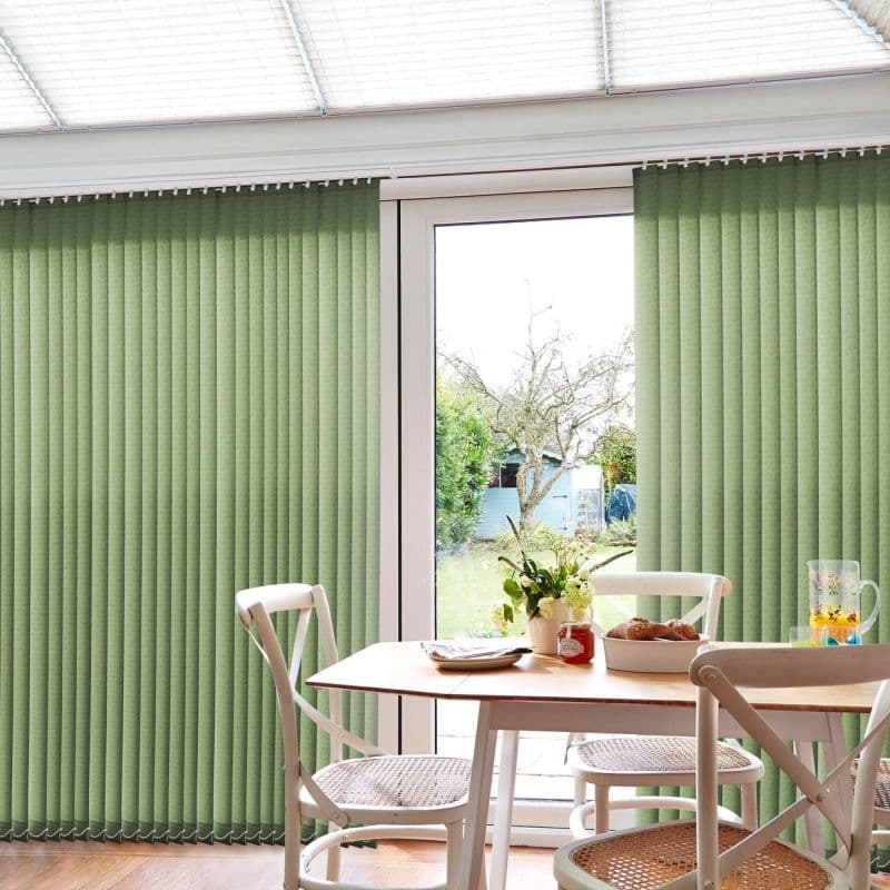 Green ribbed vertical blinds, floor-to-ceiling on a track, parted centrally to reveal a glass patio door; sunlit dining area with wooden table, white chairs, flowers, pastries, and garden view.