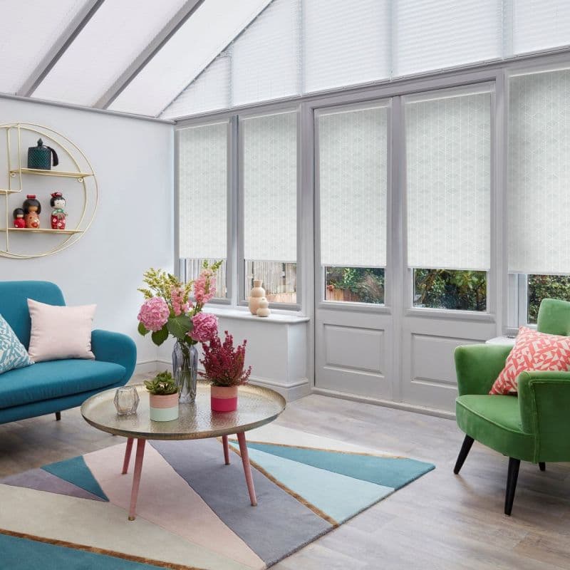 White patterned roller blinds on tall glass doors and windows, partially lowered and filtering daylight; contemporary conservatory-style living room with pastel sofa, green armchair, coffee table and flowers.
