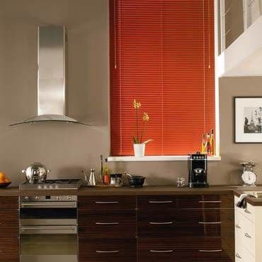 Red horizontal blinds, fully lowered over a tall window, narrow slats and pull cords; closed, filtering light; modern kitchen with stainless hood, glossy dark-wood cabinets, countertop orchid and coffee maker.