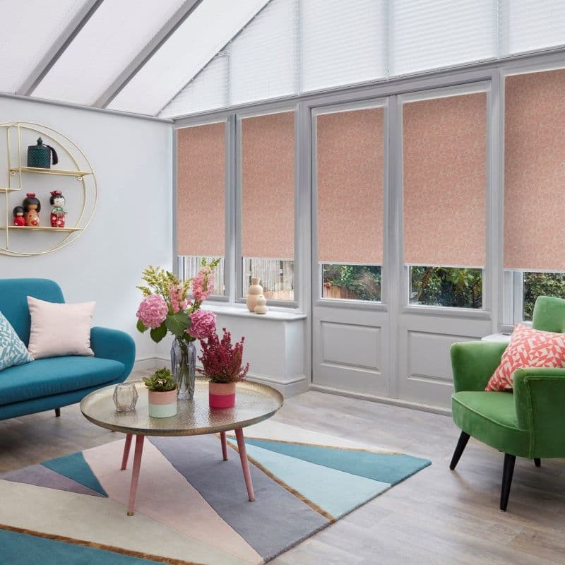Patterned pink roller blinds, lowered across four tall windows and glass door, swirled print; filtering daylight and obscuring most view; bright conservatory with slanted glass roof, colorful seating and flowers.