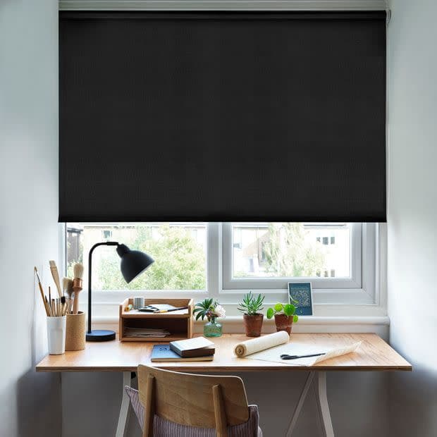 Black roller blind lowered over a double-pane window, mostly closed and blocking light; beneath it, a wooden desk with lamp, art supplies and potted succulents in a bright home study.