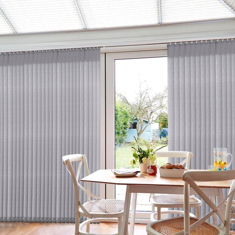 Grey vertical blinds on a track, full-height slatted panels parted at the center over a glass sliding door, partially open filtering daylight; small dining table, chairs, vase, garden outside.