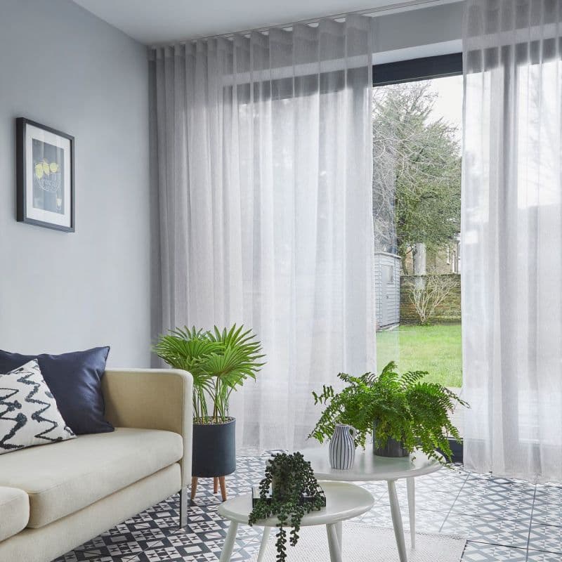 Sheer floor-to-ceiling grey curtains with soft pleats, drawn across a sliding glass door, gently filtering daylight; living room with sofa, patterned tile floor, potted plants, and a grassy backyard.
