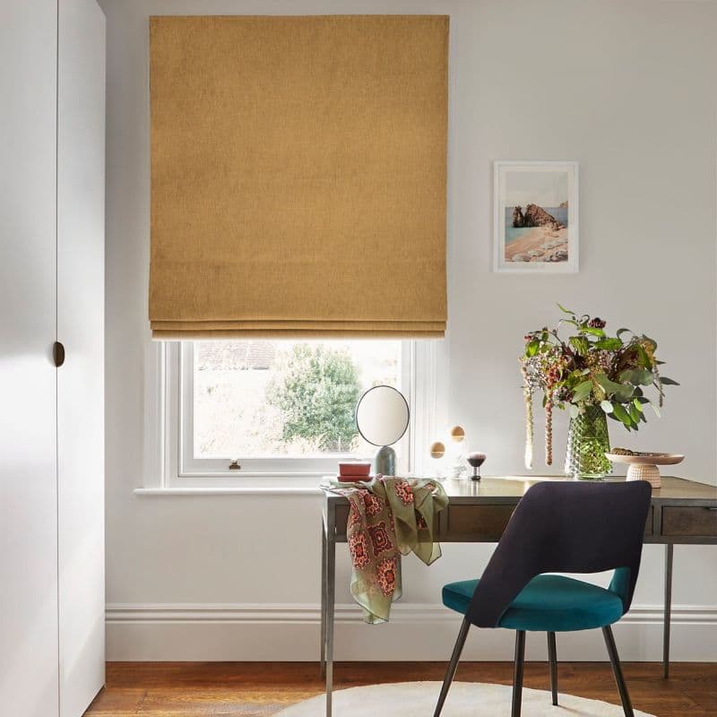 A mustard-colored fabric Roman blind, lowered with horizontal folds, filtering soft daylight into a small home office; wooden desk beneath, teal chair, mirror, vase of foliage and a garden glimpse outside.