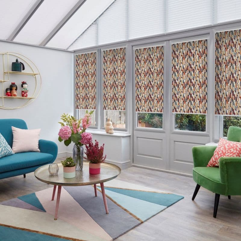 Patterned roller blinds across five tall conservatory windows/doors, lowered mid-height with geometric multicolour print, filtering daylight into a bright sunroom furnished with colorful sofas, rug, and coffee table.