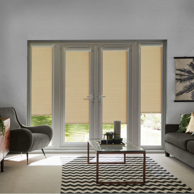 Beige roller blinds covering four-panel French doors and side panels, lowered to mid-height filtering sunlight; modern living room with grey armchairs, glass coffee table on chevron rug, glimpse of lawn.