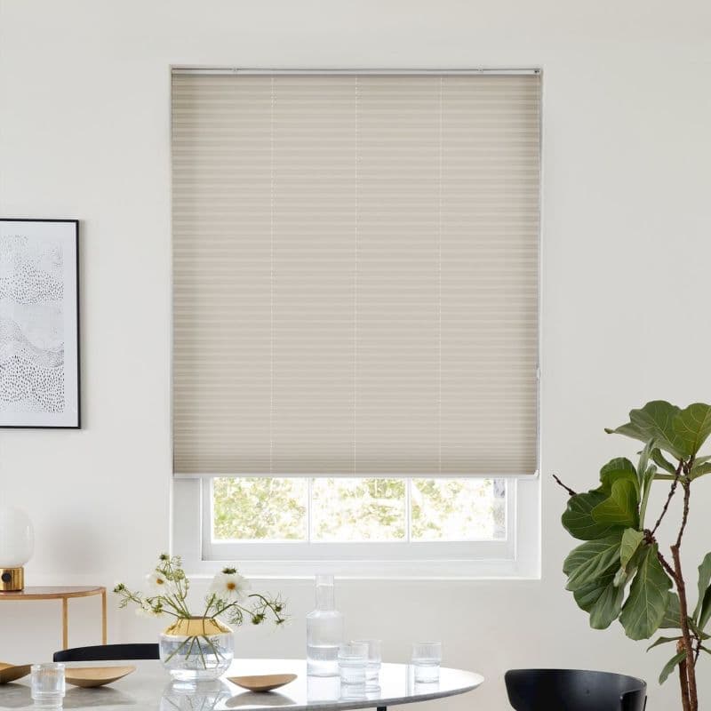 Beige pleated cellular blind lowered, with horizontal ribs and faint vertical cord lines, filtering soft daylight; over a bright minimalist dining nook with round table, vase, glassware and fiddle‑leaf fig.