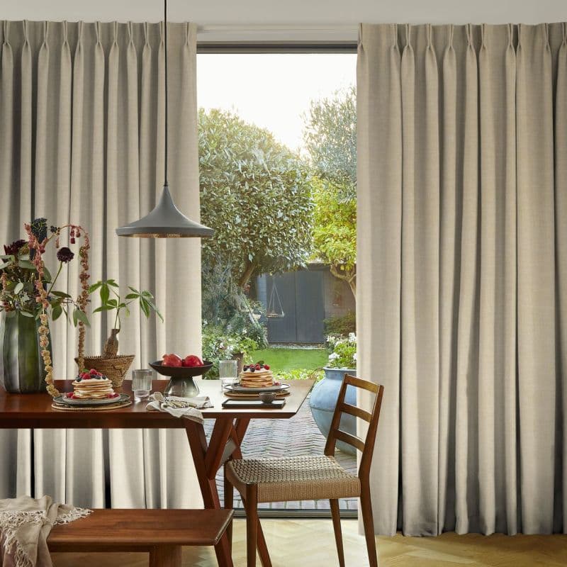 Beige pleated floor-to-ceiling curtains, mostly closed with a central gap; lightly filtering daylight and framing a glass door. Context: dining table with pendant lamp and wooden chairs, sunlit garden beyond.