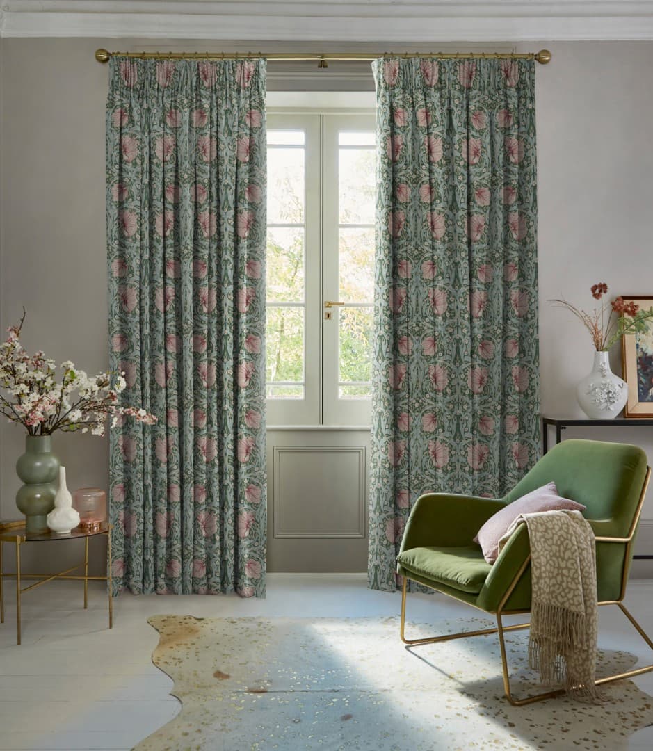 Full-length pleated floral curtains (green with pink motif) hang from a brass rod, partly drawn, filtering daylight through French doors into a softly lit room with a green armchair.