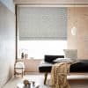 Patterned grey Roman blind, lowered with ikat motif, filters bright daylight over a window seat in a minimalist room with pale wood panels, a pendant light, cushions and a throw.
