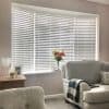 White horizontal Venetian blinds across a three-panel bay window, slats partially tilted letting soft daylight filter in; living room with gray upholstered armchairs, side table, vase of flowers on the sill.