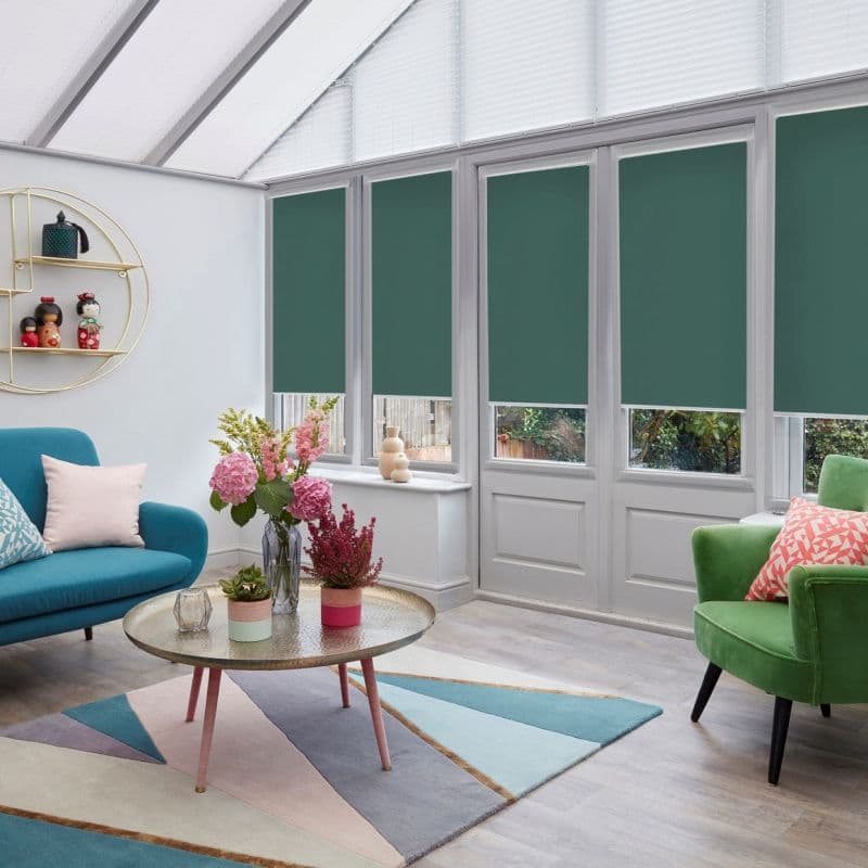 Roller blinds (dark teal) lowered across tall conservatory windows, blocking light; modern sunroom with teal sofa, green armchair, round coffee table, flowers and partial garden view.
