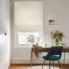 A beige fabric Roman blind, lowered with neat folds, filtering soft daylight through the window above a desk in a neutral home office with a teal chair and vase.