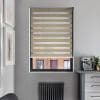 Beige zebra roller blind with alternating opaque and sheer horizontal bands, lowered across the window; filtering daylight and partially revealing a brick building outside; modern kitchen corner with coffee machine.