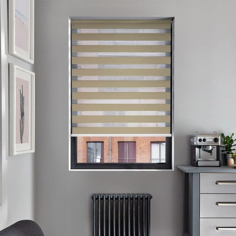 Beige zebra roller blind with alternating opaque and sheer horizontal bands, lowered across the window; filtering daylight and partially revealing a brick building outside; modern kitchen corner with coffee machine.