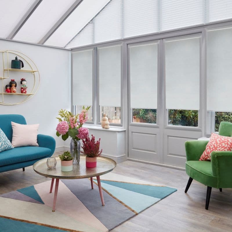 White roller blinds covering tall conservatory windows and glazed doors, lowered to mid-height and softly filtering daylight; modern sunroom with teal sofa, green armchair, round coffee table and flowers.