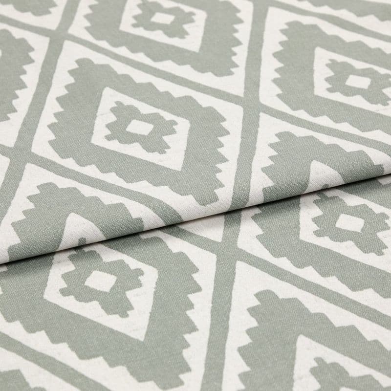 Folded woven fabric featuring repeating jagged-diamond, Aztec-like motif in sage green and off-white — layered across itself, lying flat in a soft, evenly lit close-up highlighting weave texture.